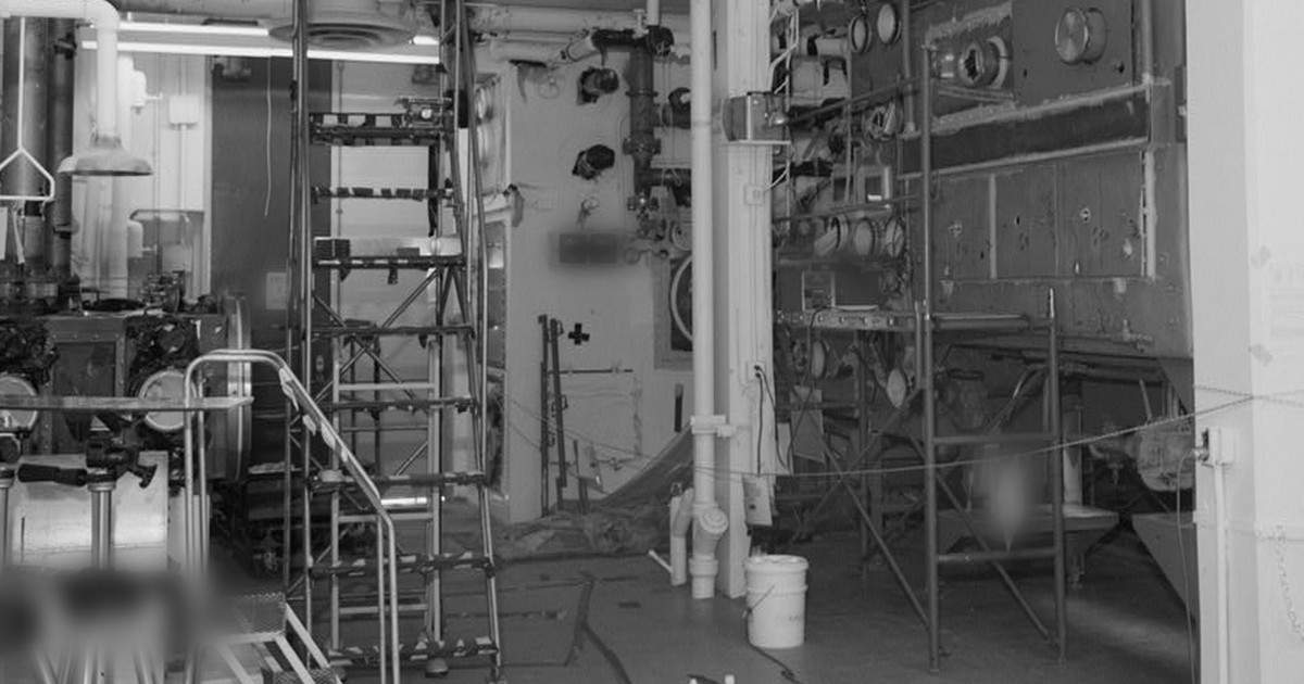 Sala industrial em preto e branco com caixas de luvas metálicas, escadas e tubulações