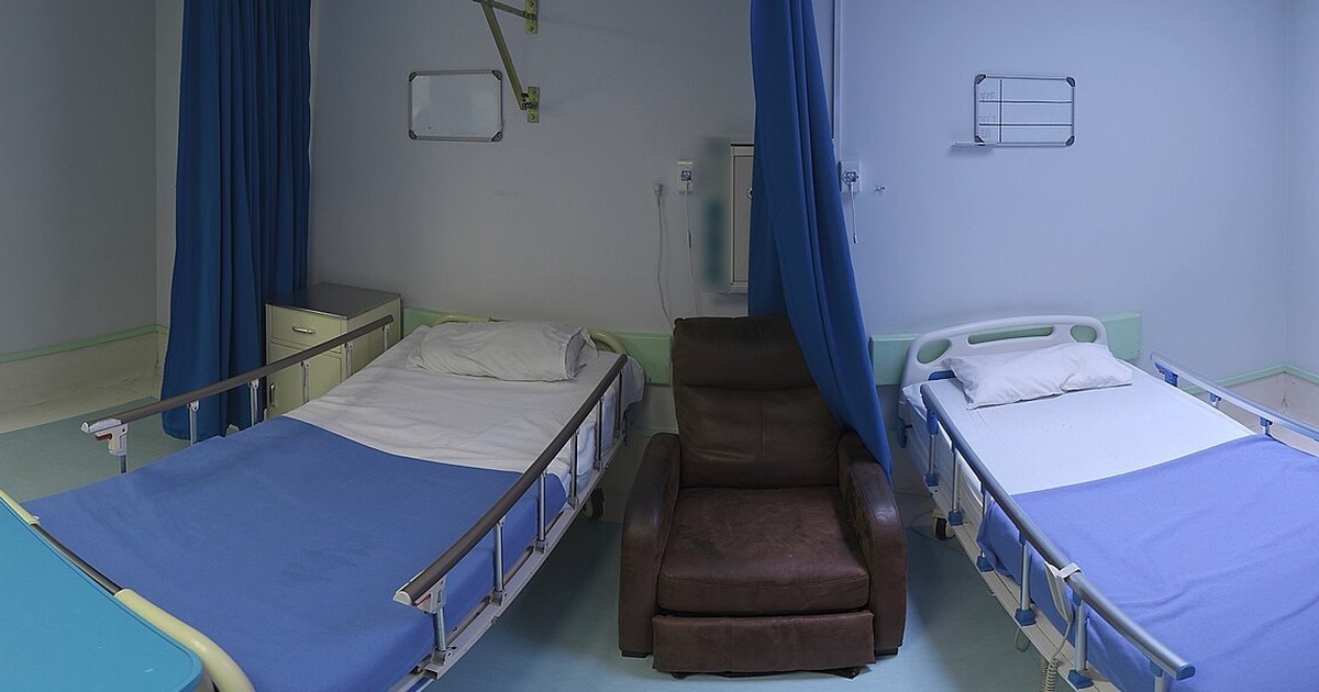 Enfermaria hospitalar vazia com dois leitos de lençóis azuis, cortinas azuis e uma poltrona marrom entre eles