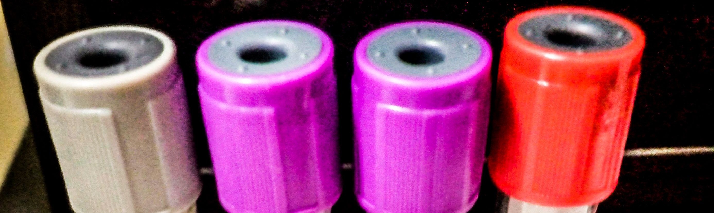 Tampas coloridas de quatro tubos de coleta de sangue lado a lado, em cinza, roxo e vermelho, sobre fundo escuro