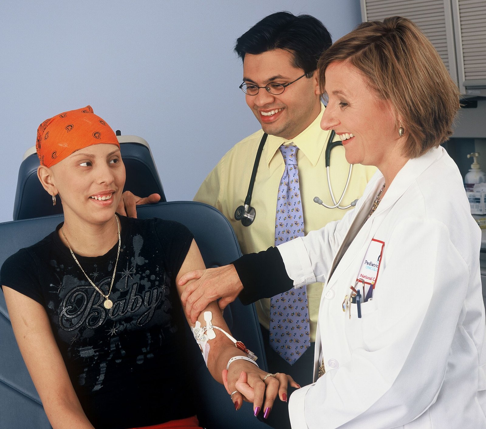 A female doctor in a white coat and a male doctor in a yellow shirt, tie and stethoscope around his neck smile next to a young woman with an orange headscarf, seated in a blue treatment chair, with an intravenous line attached to her forearm.
