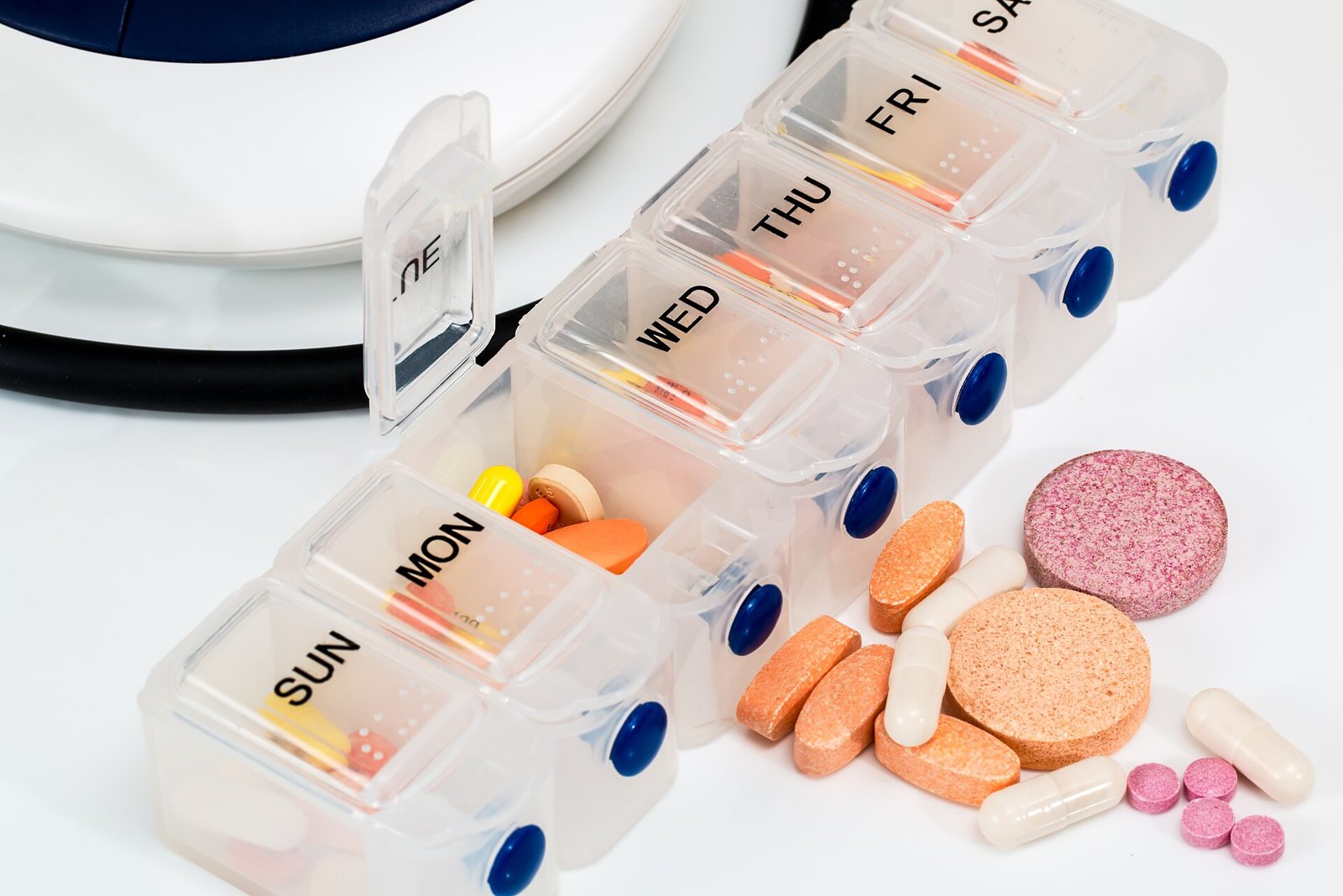 A transparent weekly pill organizer with blue lids and the days of the week in English, with colored pills in some compartments and, next to it, white capsules, orange tablets and pink effervescent tablets on a white surface.