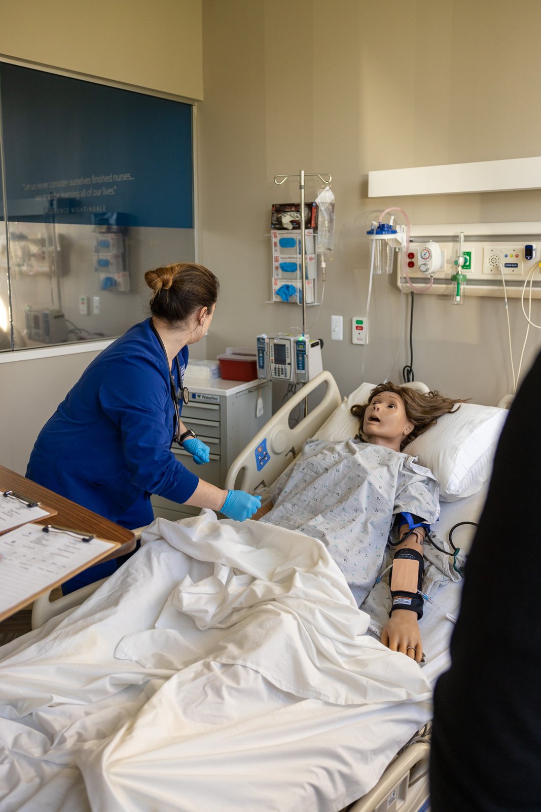 A nursing student in a navy blue uniform and blue gloves examines a simulation mannequin lying in a hospital bed, wearing a patterned gown and a blood pressure cuff on the arm, in a room with an IV pole, infusion pumps and a gas outlet rail on the wall.
