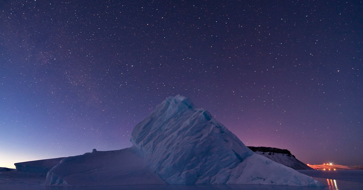 Iceberg branco na baía de North Star, na Groenlândia, sob um céu noturno estrelado, com o mar escuro e calmo à frente.