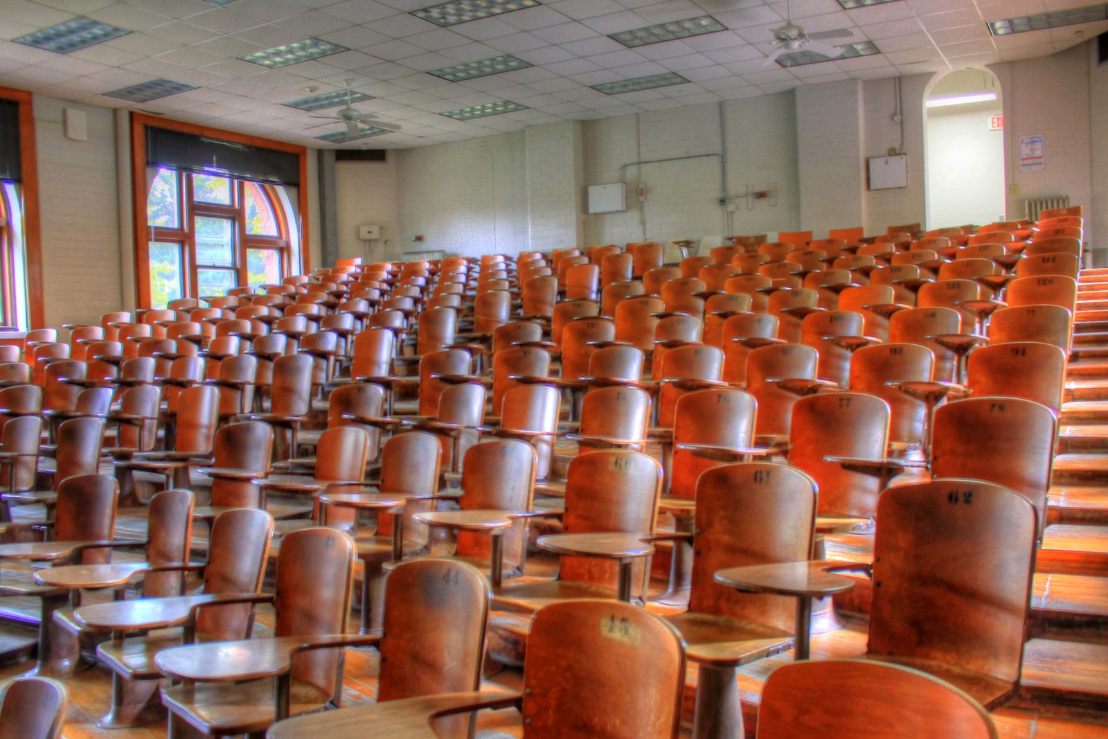 Grande auditório universitário vazio, com dezenas de fileiras de cadeiras de madeira com braço de apoio, voltadas para a frente da sala.