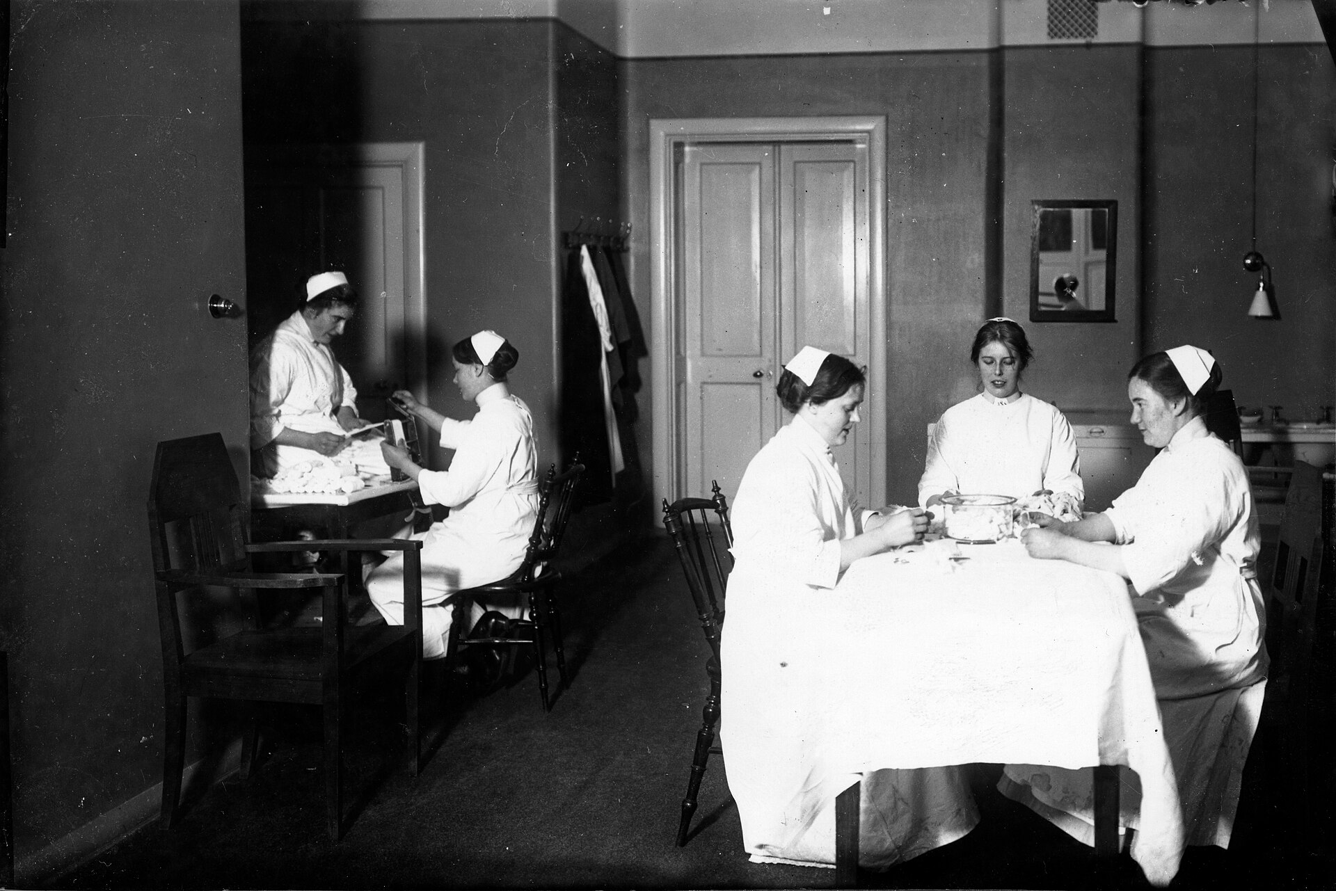Photo ancienne en noir et blanc d'une salle d'hôpital où cinq infirmières en coiffe blanche, assises autour d'une table recouverte d'une nappe, enroulent des bandes de gaze sorties d'un bol en verre