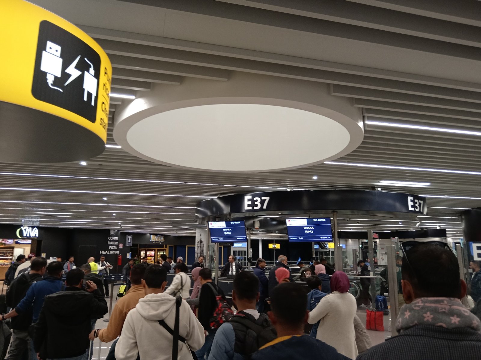 Hall d'embarquement d'un aéroport avec des passagers en file, des panneaux de porte et un plafond avec un luminaire circulaire.