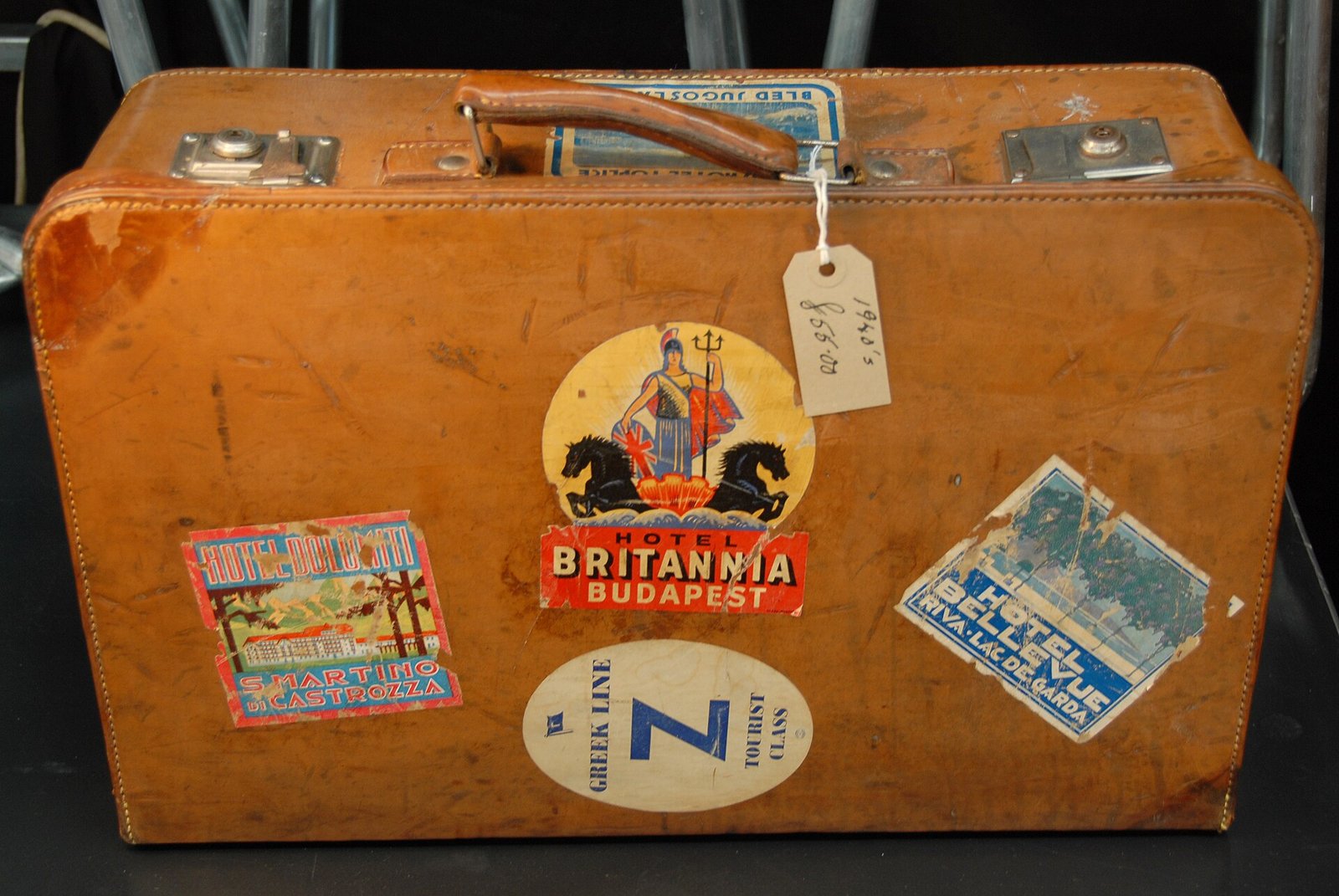 Vieille valise en cuir marron, avec poignée, fermoirs métalliques et autocollants d'hôtels collés sur le devant, une étiquette en papier attachée à la poignée, sur une surface sombre.