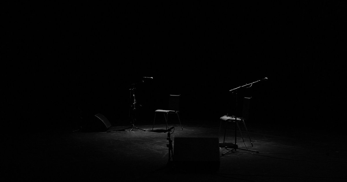 Escenario oscuro en blanco y negro, iluminado solo en el centro, con dos sillas vacías, dos pedestales de micrófono y altavoces en el suelo.