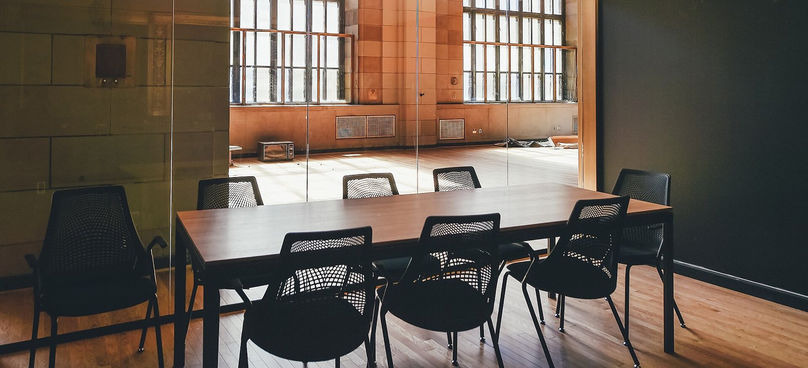 A meeting room with a long wooden table and black chairs with open backs, seen through a glass wall; in the background, a large hall with tall windows and a sunlit wooden floor.