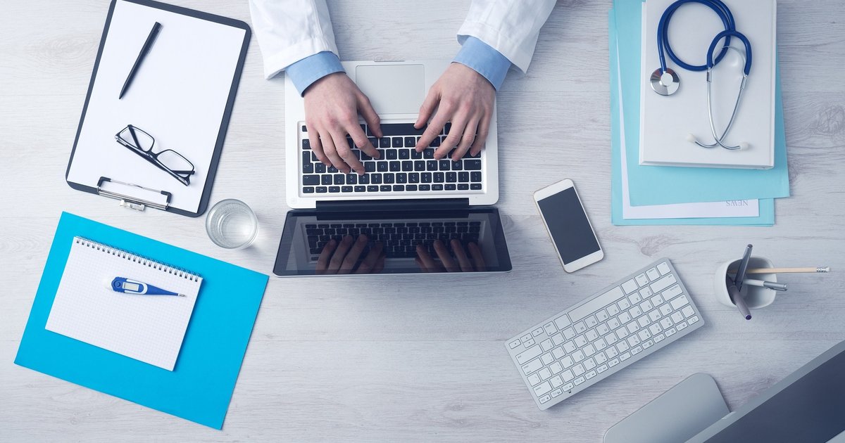 Mesa de trabalho médica vista de cima com notebook, estetoscópio, óculos, prancheta e termômetro