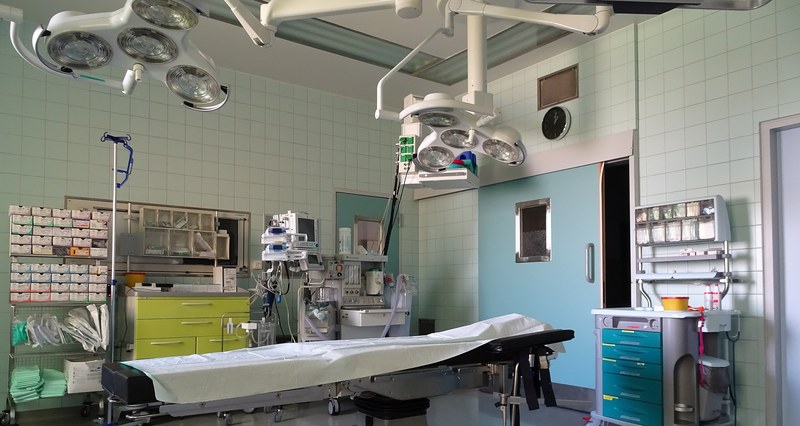 Sala cirúrgica atual com mesa de operação coberta, focos de luz no teto, carrinho de anestesia e armários de material