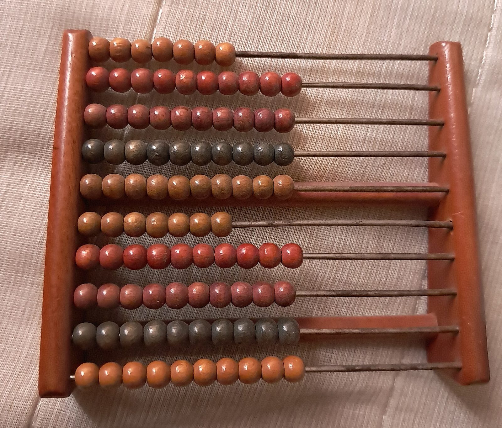 Ábaco pequeno de madeira com dez fileiras de contas vermelhas, laranja e verde-acinzentadas em hastes de metal, sobre um tecido bege