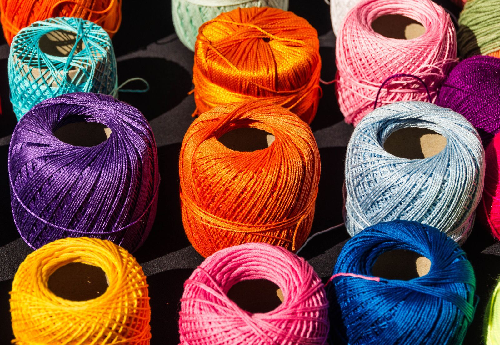 Novelos de linha de algodão em laranja, roxo, rosa, azul-claro, azul, amarelo e turquesa, enfileirados sobre um fundo escuro