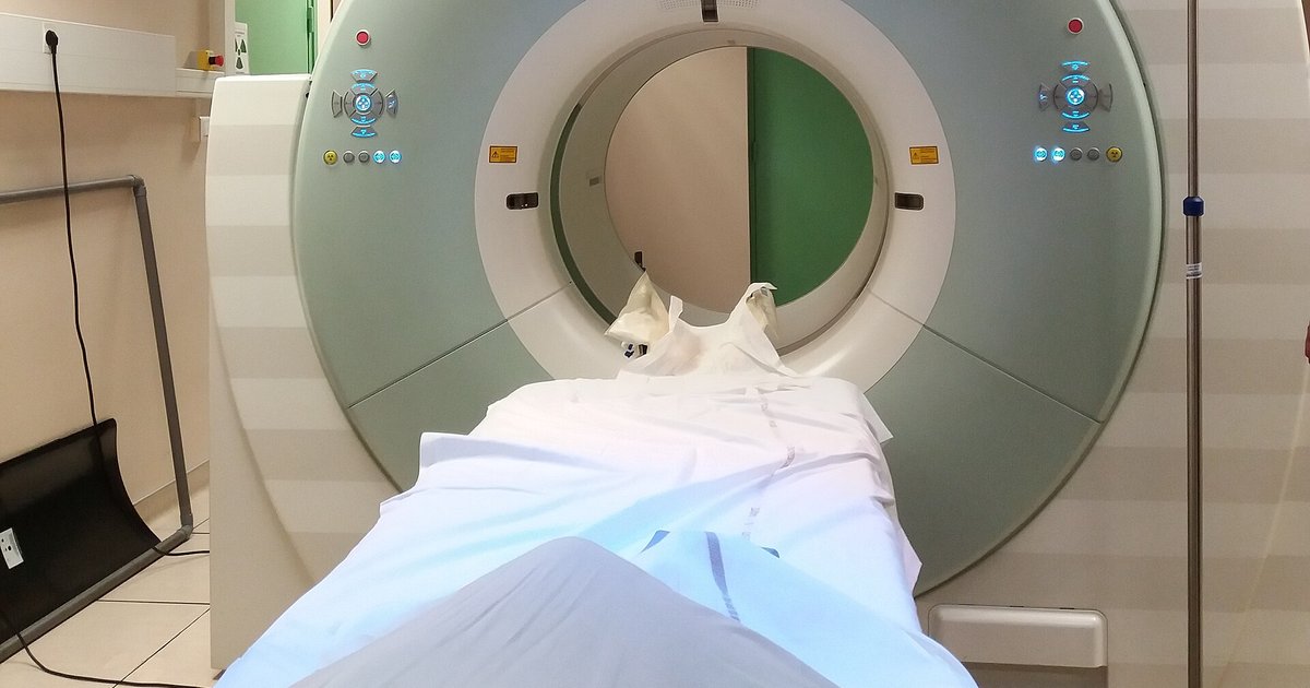 Aparelho de PET com tomografia visto de frente, com a maca coberta por lençol branco entrando no anel do equipamento, em sala de hospital