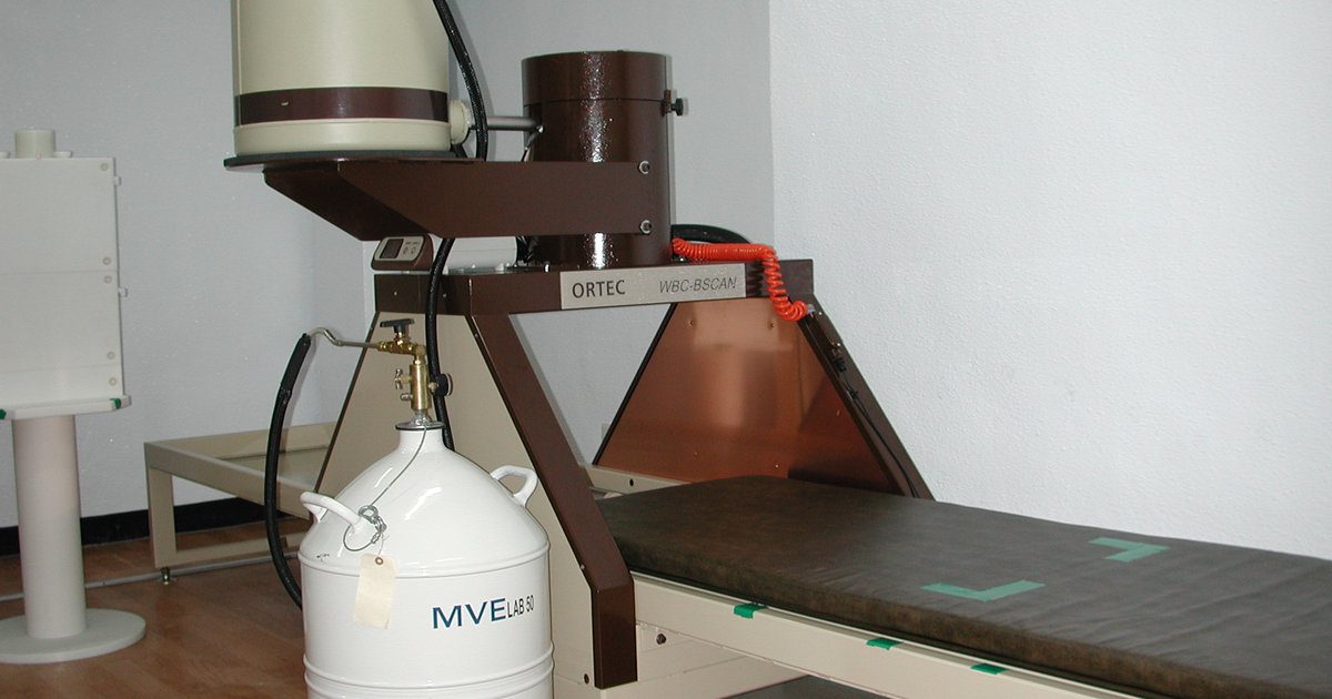 Sala clínica com maca estreita sob um detector suspenso em braço metálico e cilindros de nitrogênio líquido ao lado