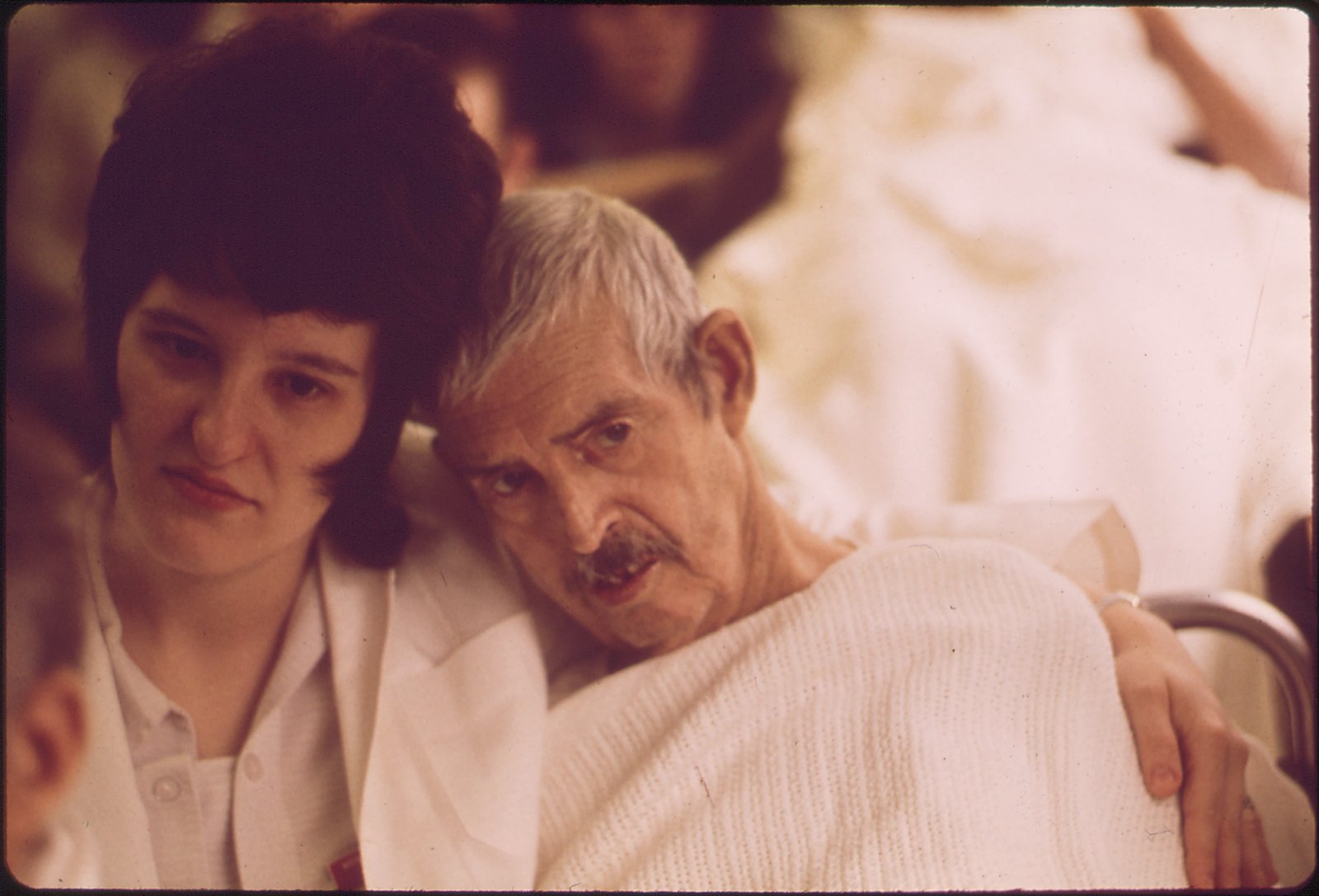 Photographie ancienne aux tons chauds d'une jeune femme aux cheveux foncés et en blouse blanche qui entoure de son bras les épaules d'un homme âgé aux cheveux blancs et à la moustache, enveloppé dans une couverture claire ; il pose la tête contre la sienne.