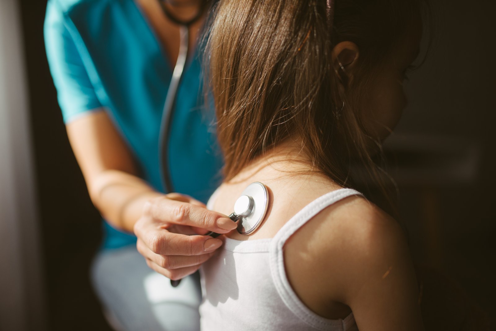 Main d'une professionnelle en uniforme bleu posant le stéthoscope sur le dos d'une fillette aux longs cheveux et en débardeur blanc, dans une salle à la lumière latérale.
