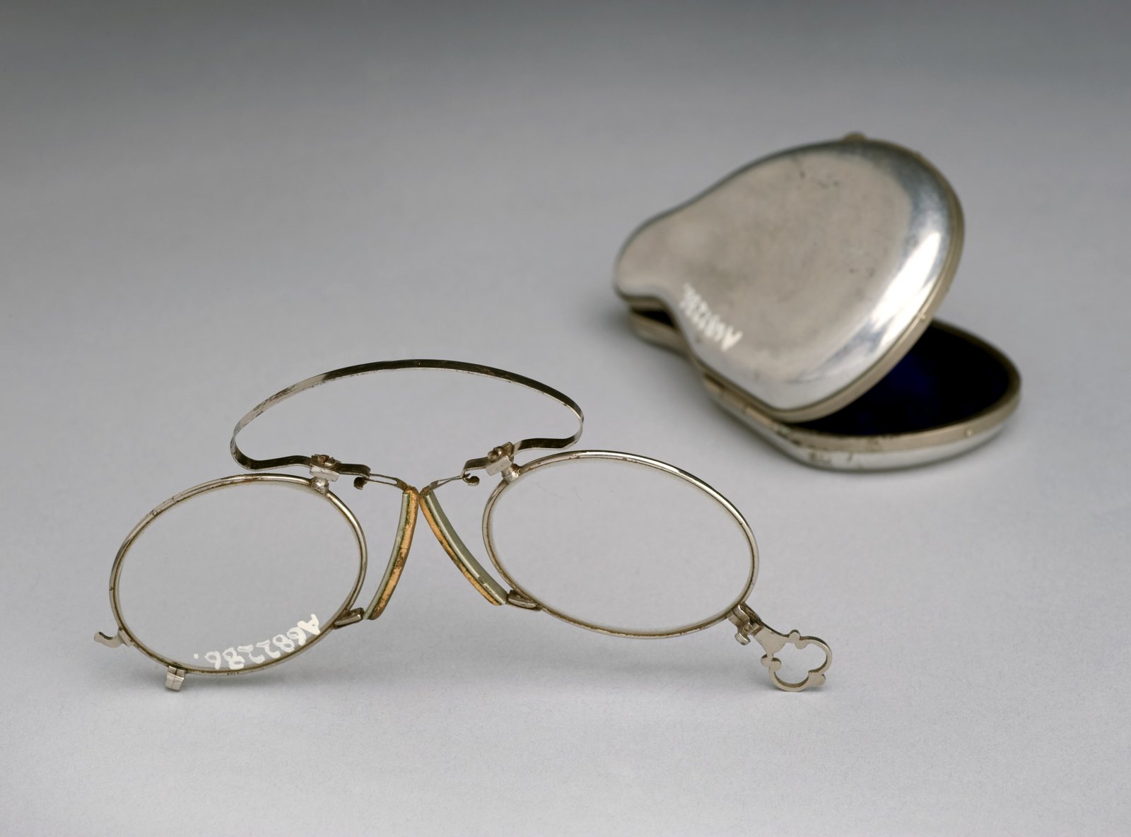 Alte Brille mit dünnem Metallrahmen und Feders teg über hellgrauem Hintergrund, mit einer kleinen, auf eine der Brillengläser gemalten Katalognummer und daneben das geöffnete Metall-Etui mit blauem Futter.