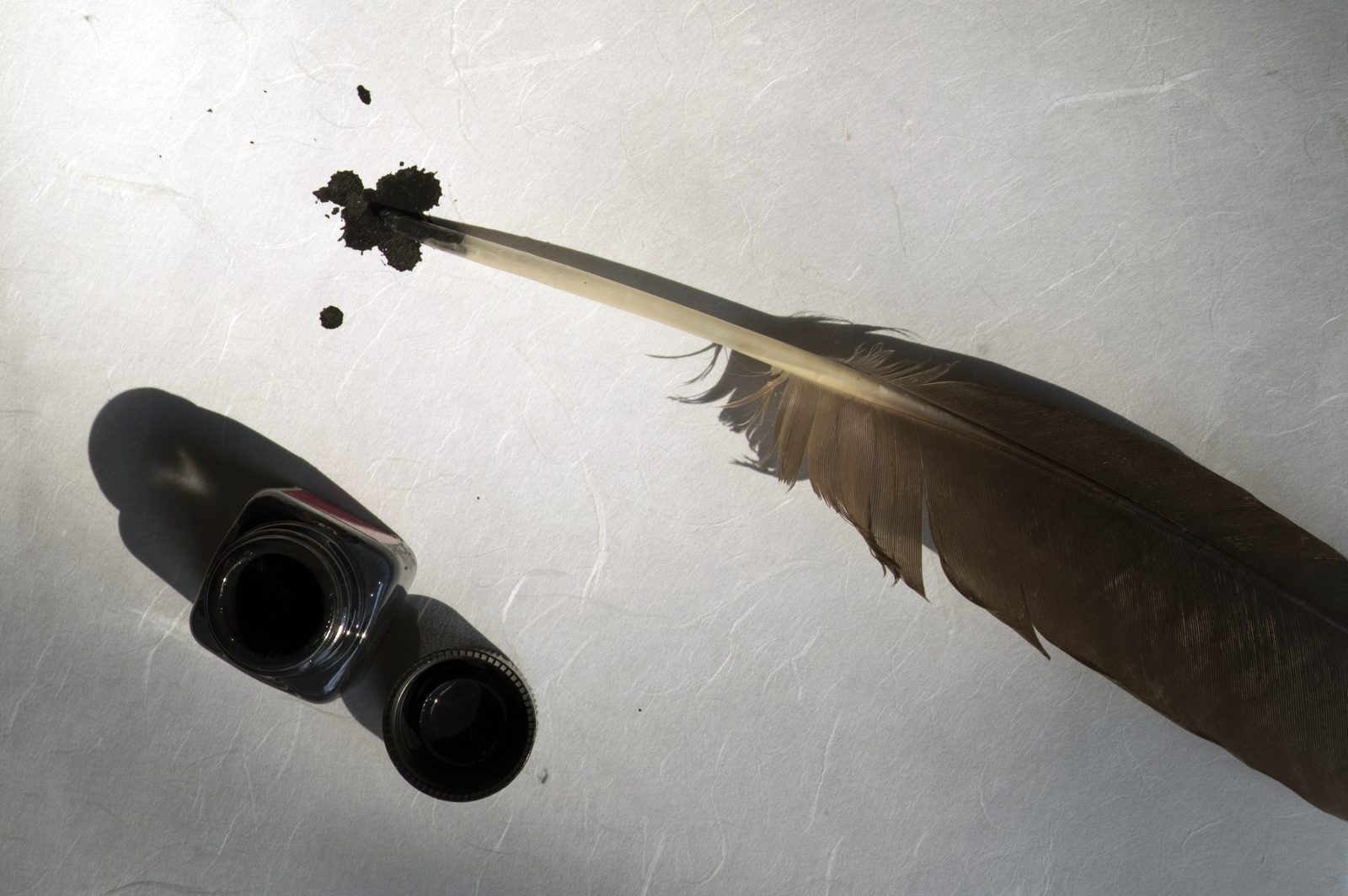 Plume d'écrire posée sur une feuille blanche, à côté d'un encrier sombre renversé, avec une tache d'encre sur le papier.