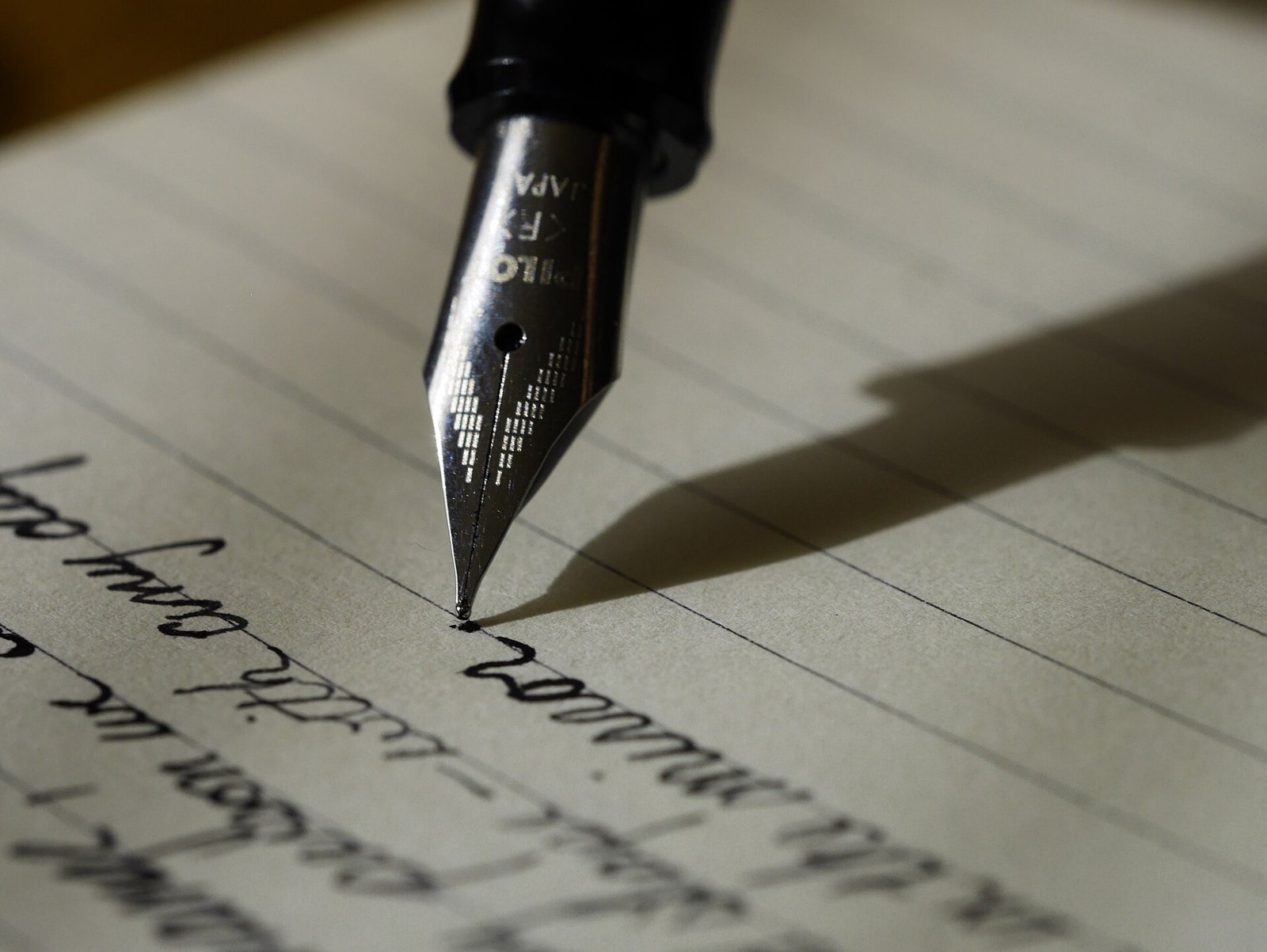 Gros plan d'un stylo-plume écrivant à la main en lettres cursives sur une feuille lignée, avec la pointe métallique au point et l'ombre du stylo sur le papier.