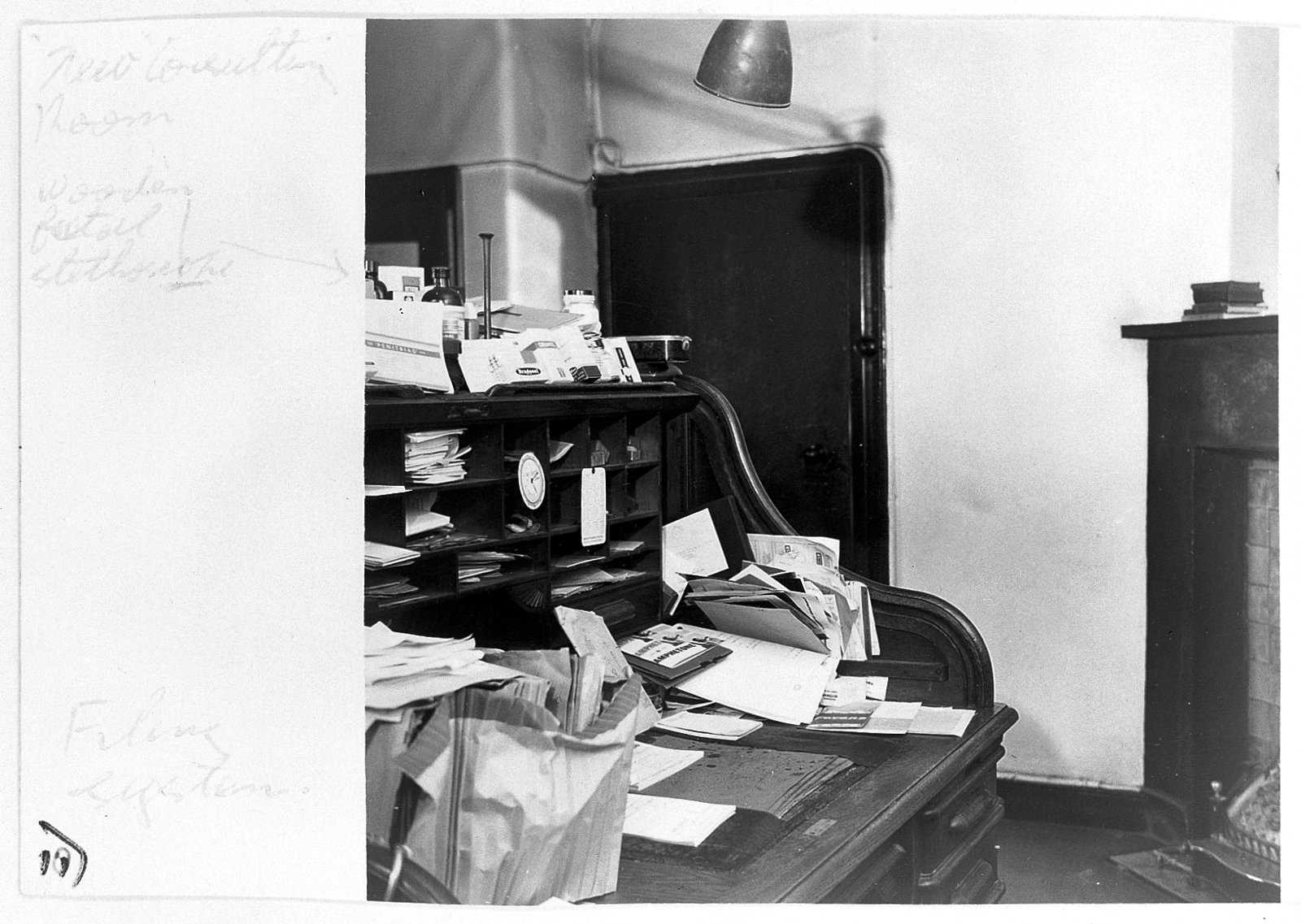 Photographie ancienne en noir et blanc d'un bureau de cabinet couvert de papiers et de fiches, avec des casiers et des flacons en hauteur, et des annotations à la main dans la marge de la photo.