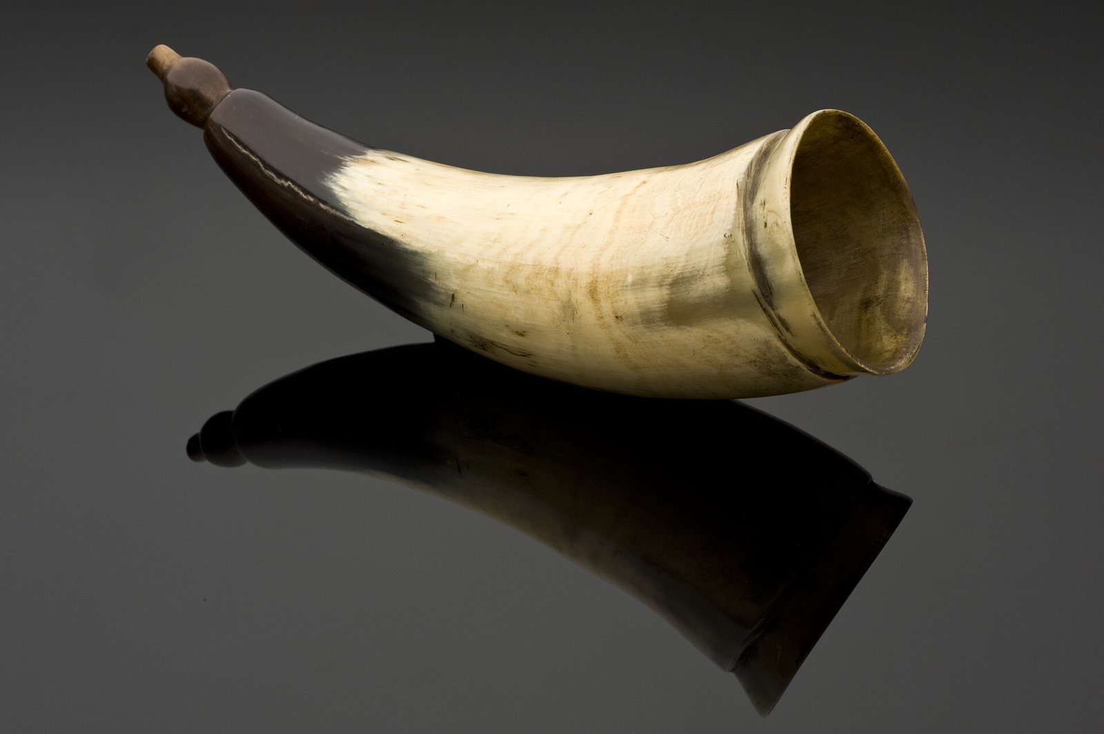 Ancien cornet acoustique en corne claire, avec l'embouchure large garnie de métal doré et l'extrémité sombre pour l'oreille, posé sur un fond noir brillant qui reflète l'objet.
