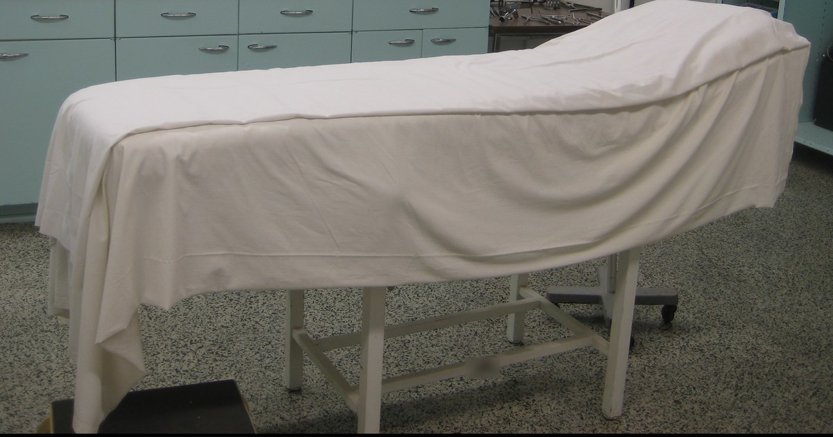 Maca hospitalar coberta por um lençol branco diante de armários verde-claros em uma sala de atendimento