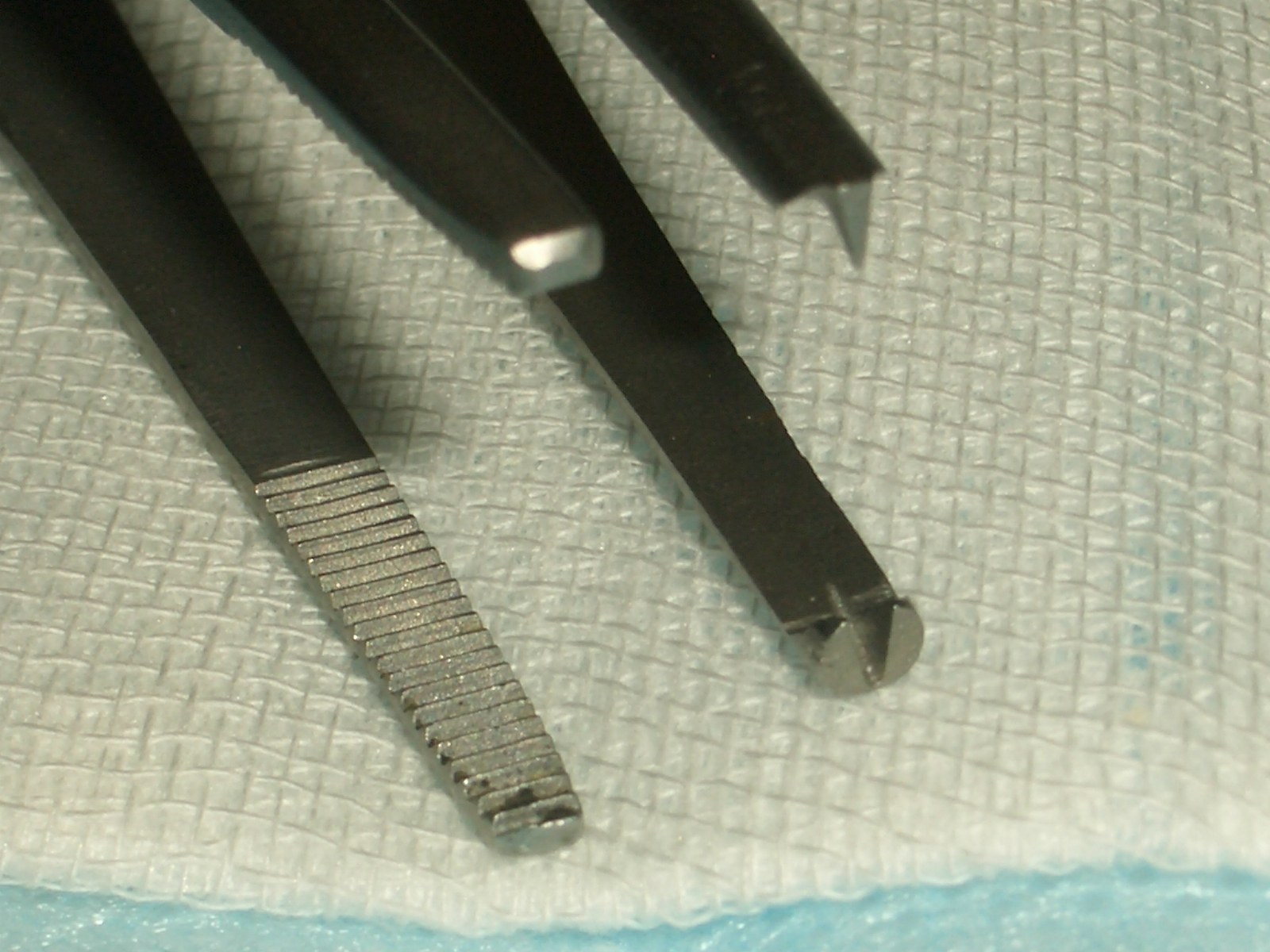 Pinças de dissecção de aço com pontas serrilhadas e uma com dentes na ponta, sobre um tecido claro