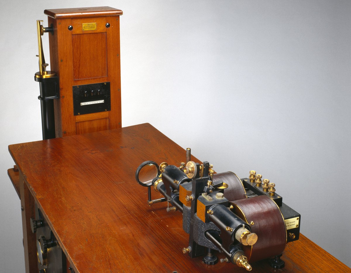 Eletrocardiógrafo histórico, com galvanômetro metálico e caixa de registro sobre mesa de madeira