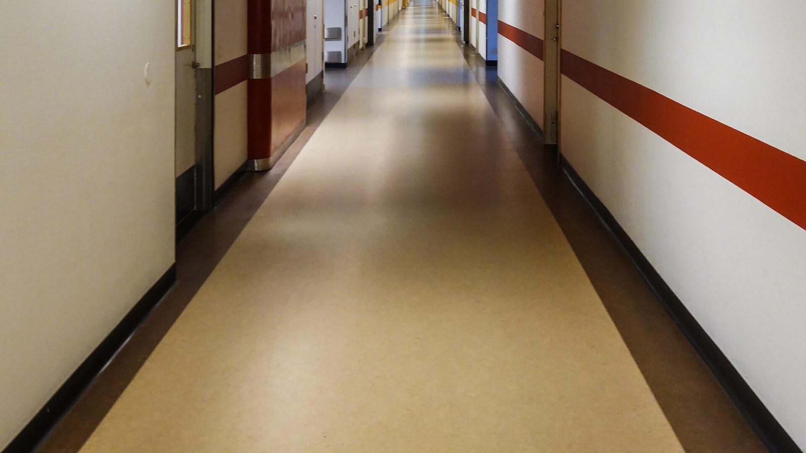 Pasillo largo y vacío de hospital, de piso beige y paredes claras con una franja roja, puertas a ambos lados y luz al fondo