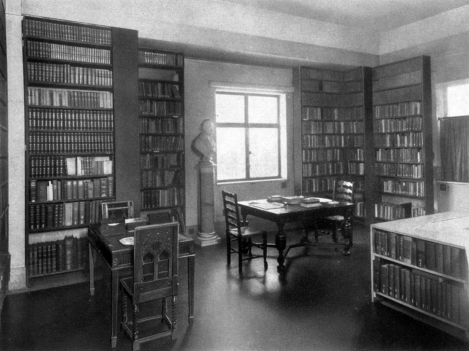 Photographie ancienne en noir et blanc d'un coin de salle de lecture d'une bibliothèque médicale, avec des rayonnages de livres jusqu'au plafond, un buste sur un piédestal, des tables avec des chaises et une fenêtre éclairant la pièce.