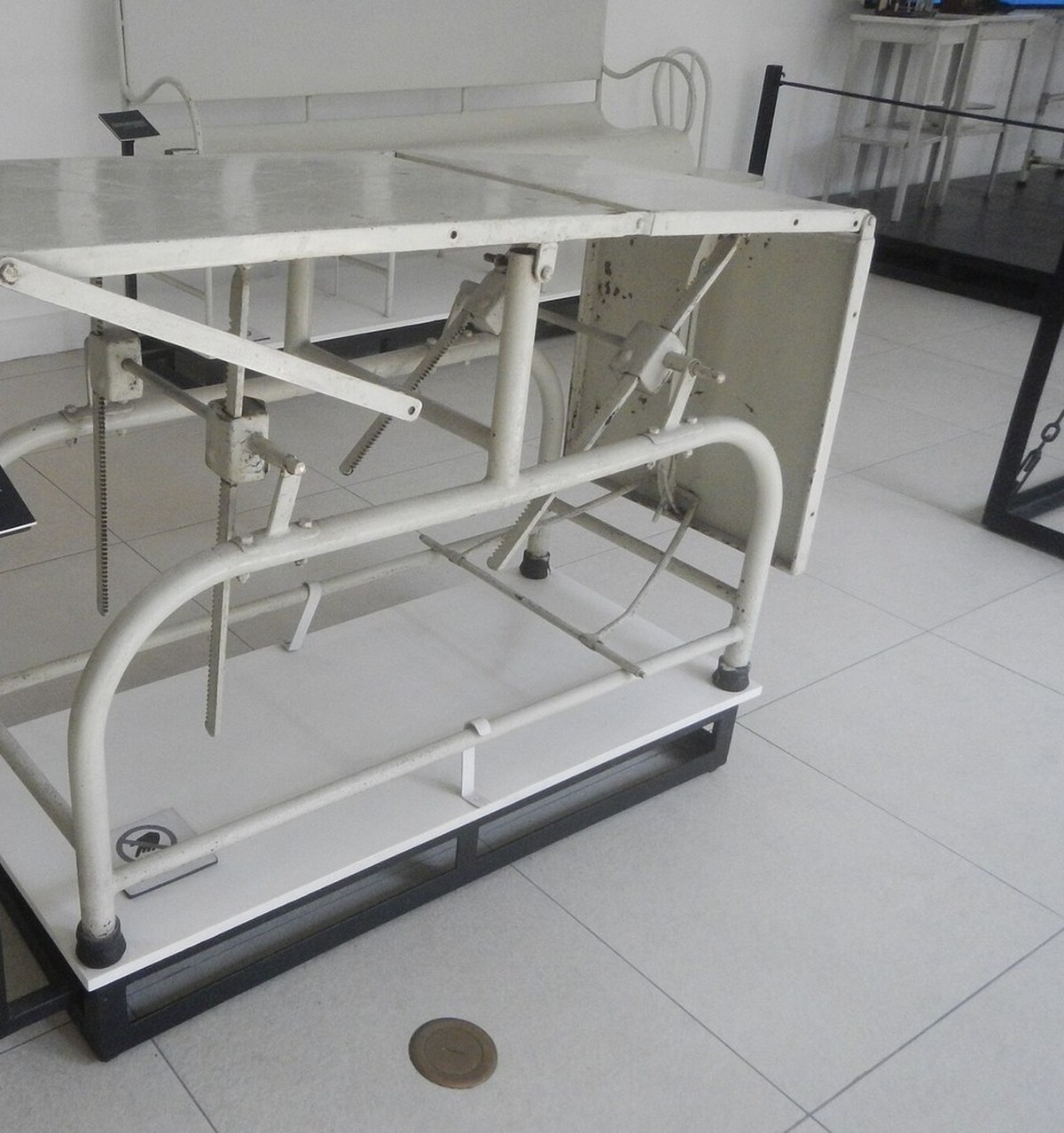 Mesa de exame antiga de metal branco, exposta em sala de museu sem pessoas
