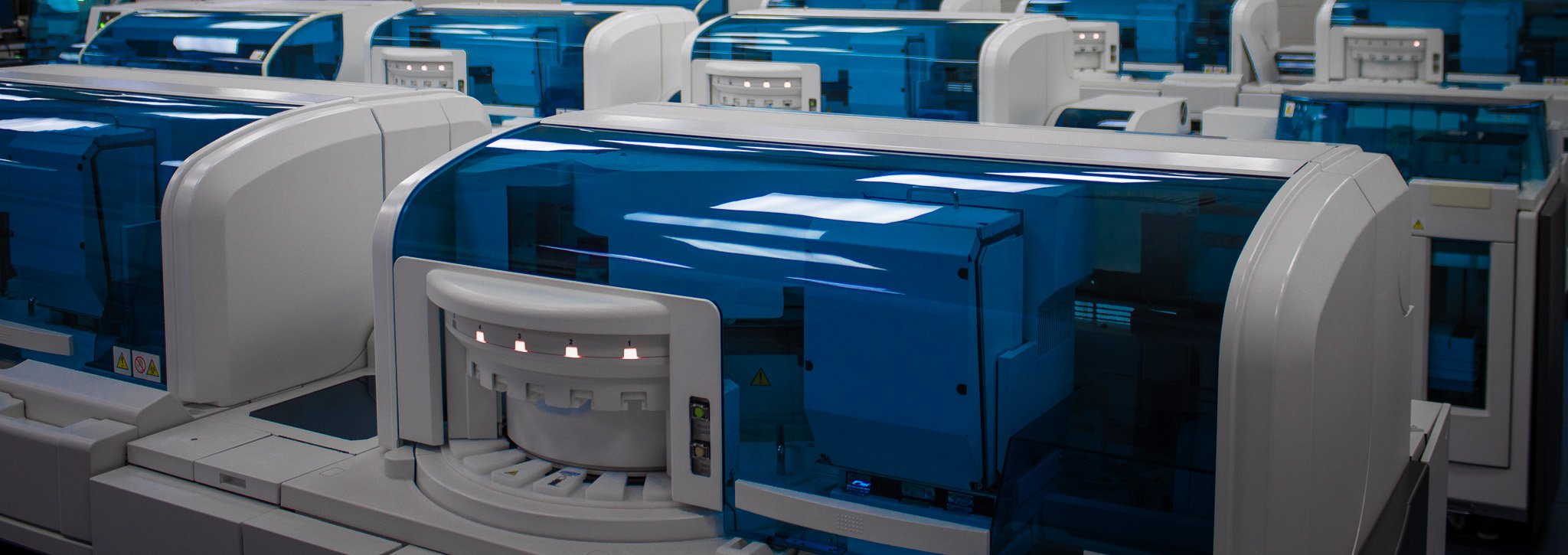 Fileiras de analisadores automáticos de laboratório clínico com tampas azuis transparentes numa sala ampla e iluminada