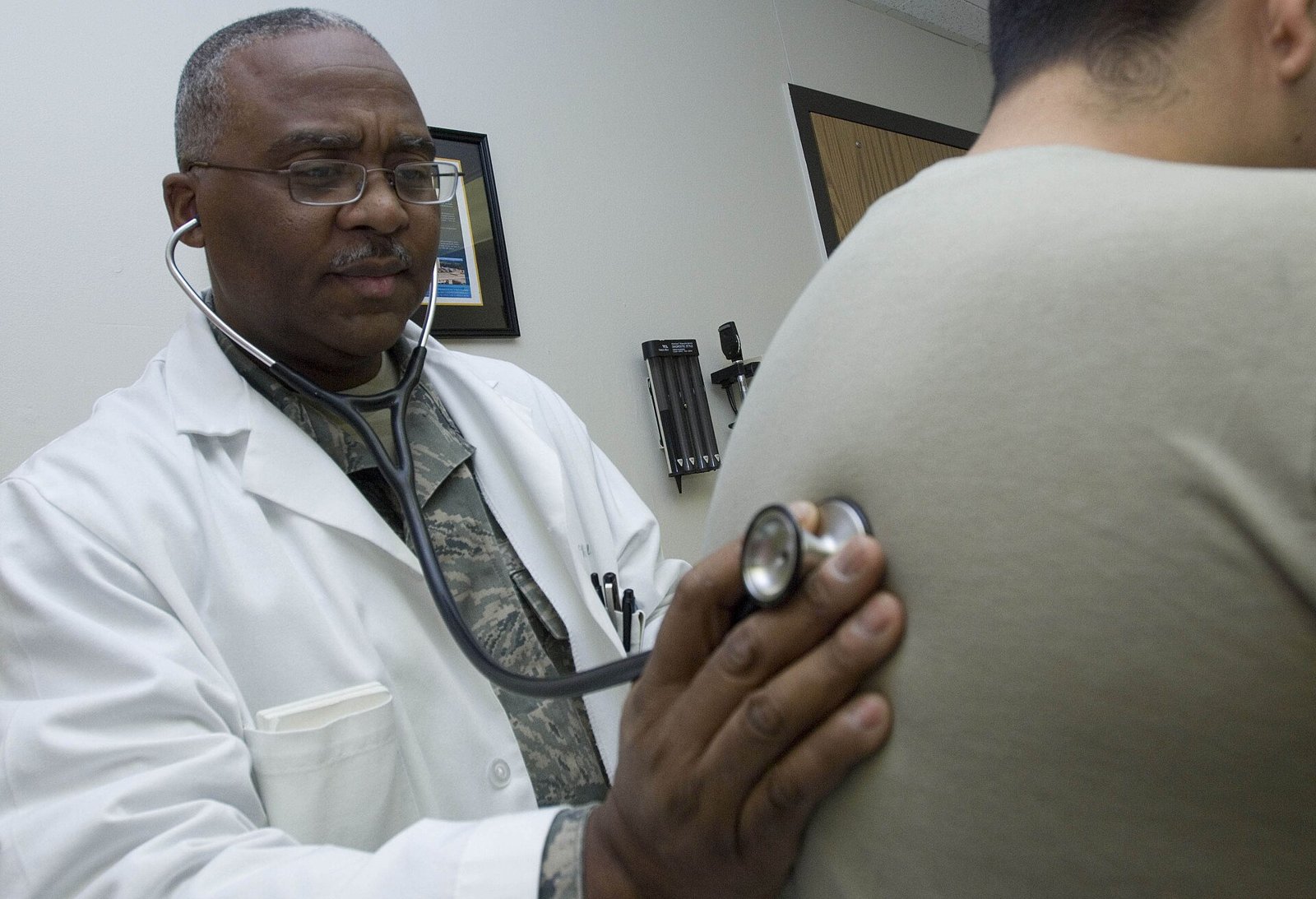 Doctor in a white coat auscultating a patient's back with a stethoscope, in an office.