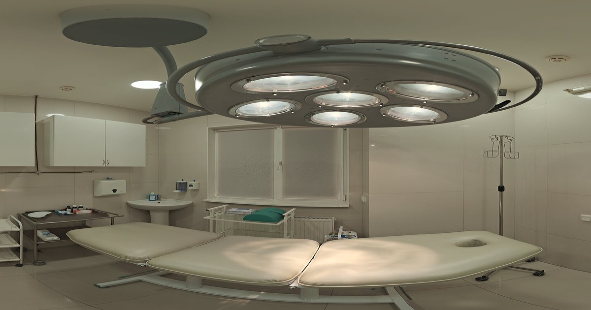 Sala de exame clara com maca acolchoada bege sob um foco cirúrgico de várias lâmpadas e um suporte de soro
