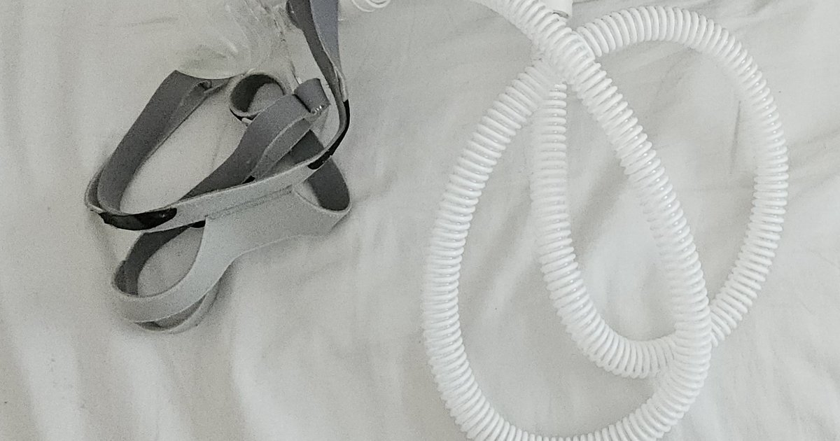 Mangueira branca corrugada de CPAP enrolada sobre lençol branco, ao lado de tiras cinza de máscara