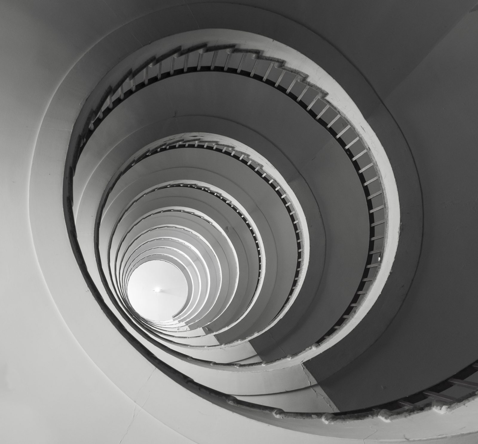 Escada em espiral vista de baixo, em preto e branco, com os degraus formando anéis concêntricos que sobem até uma claraboia.