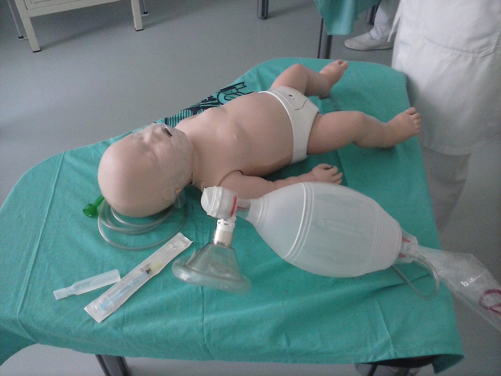 Eine Babypuppe für Reanimationstraining liegt auf einem mit grünem Tuch bedeckten Tisch, daneben ein Beatmungsbeutel mit Maske, eine Spritze und eine Sonde.