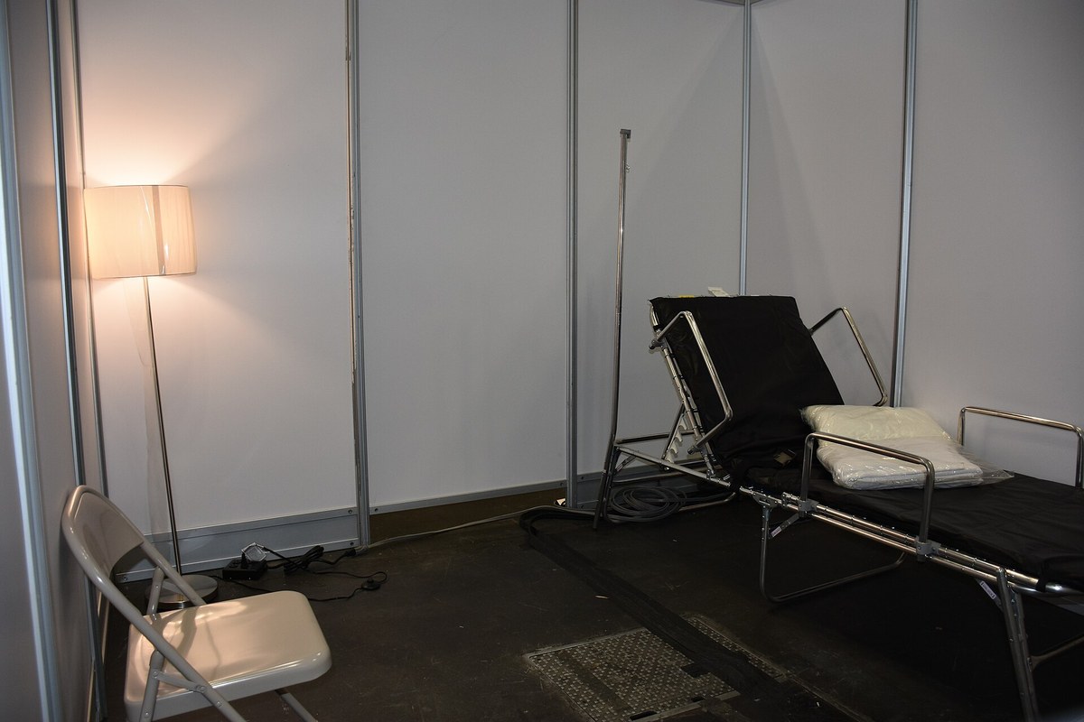 Sala de repouso com cama dobrável, cadeira e luminária, sem pessoas