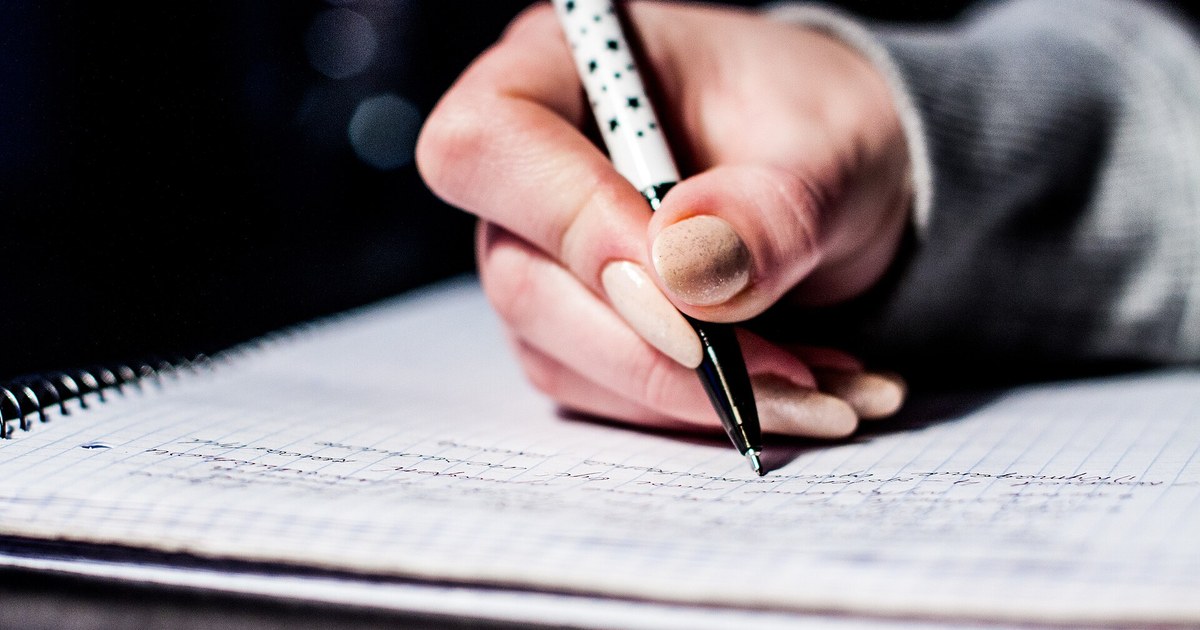 A hand with light-colored nails holding a white pen with black polka dots, writing by hand in a spiral notebook with grid paper, against a dark background