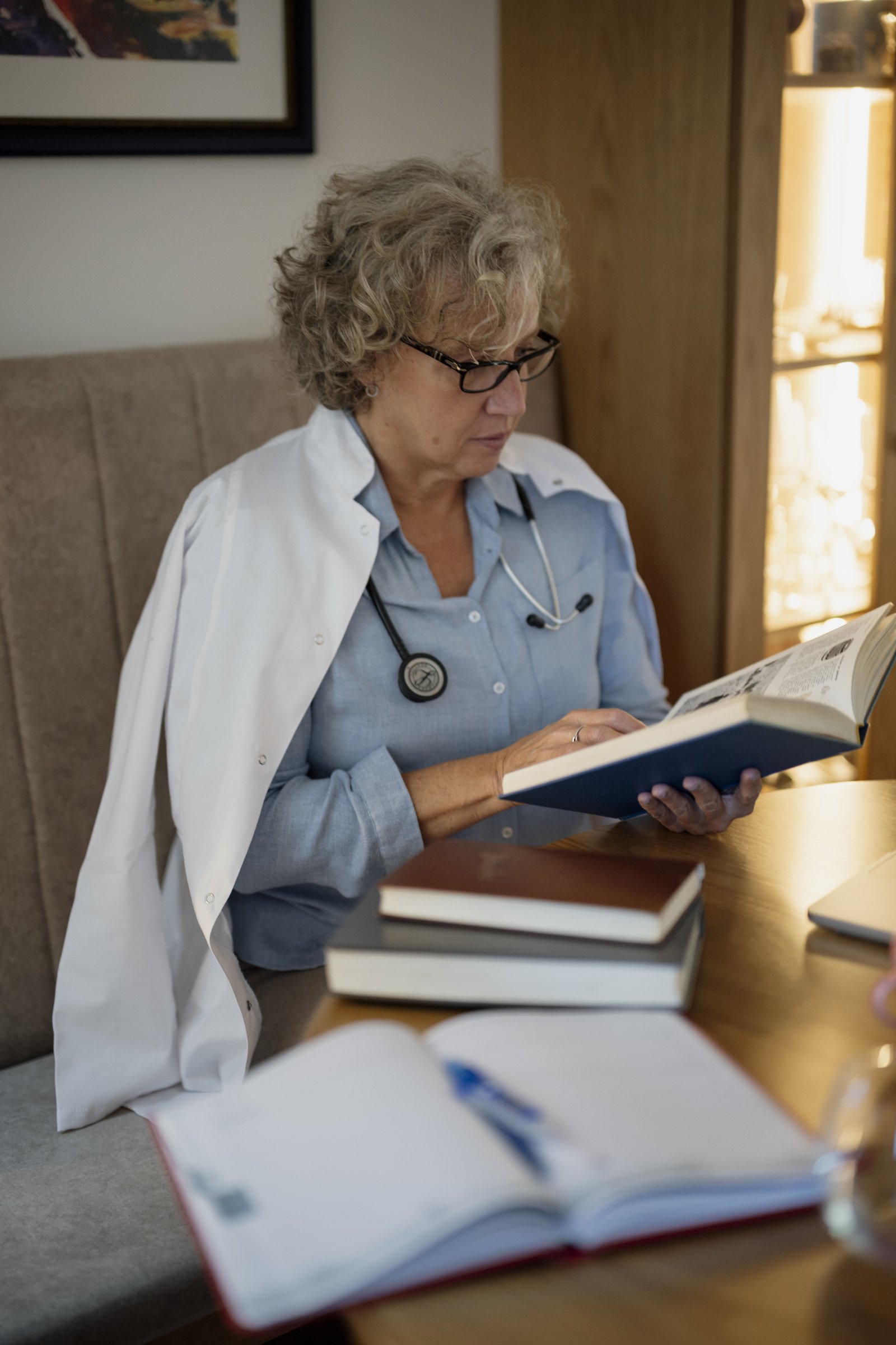 A doctor with curly gray hair and glasses, wearing a light blue shirt, a stethoscope around her neck and a white coat over her shoulders, sitting on a padded bench reading a book with a blue cover, with other books and an open notebook on the wooden table.