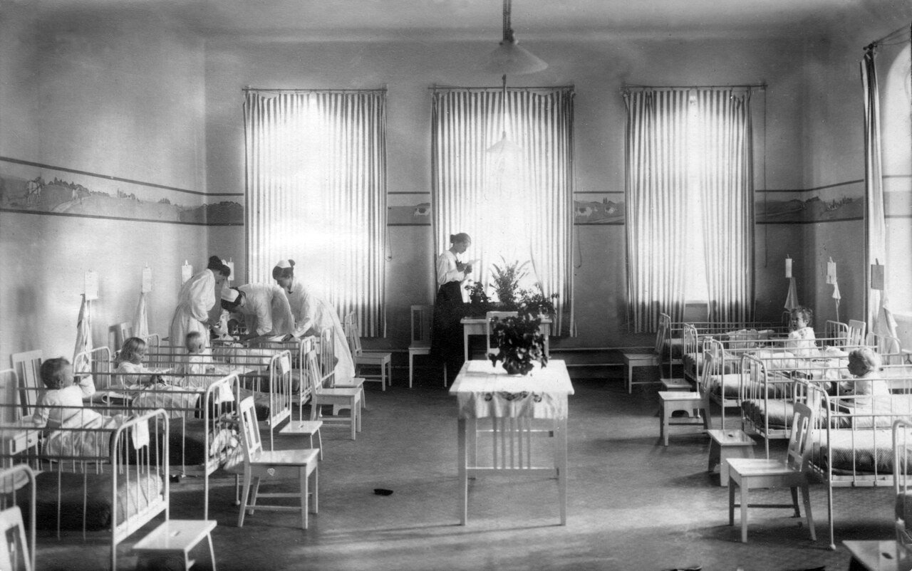 Old black and white photo of a children's ward lit by three tall windows with striped curtains: children in white barred cribs on both sides, three nurses in white leaning over a bed on the left and a fourth next to a table with potted plants in the center.