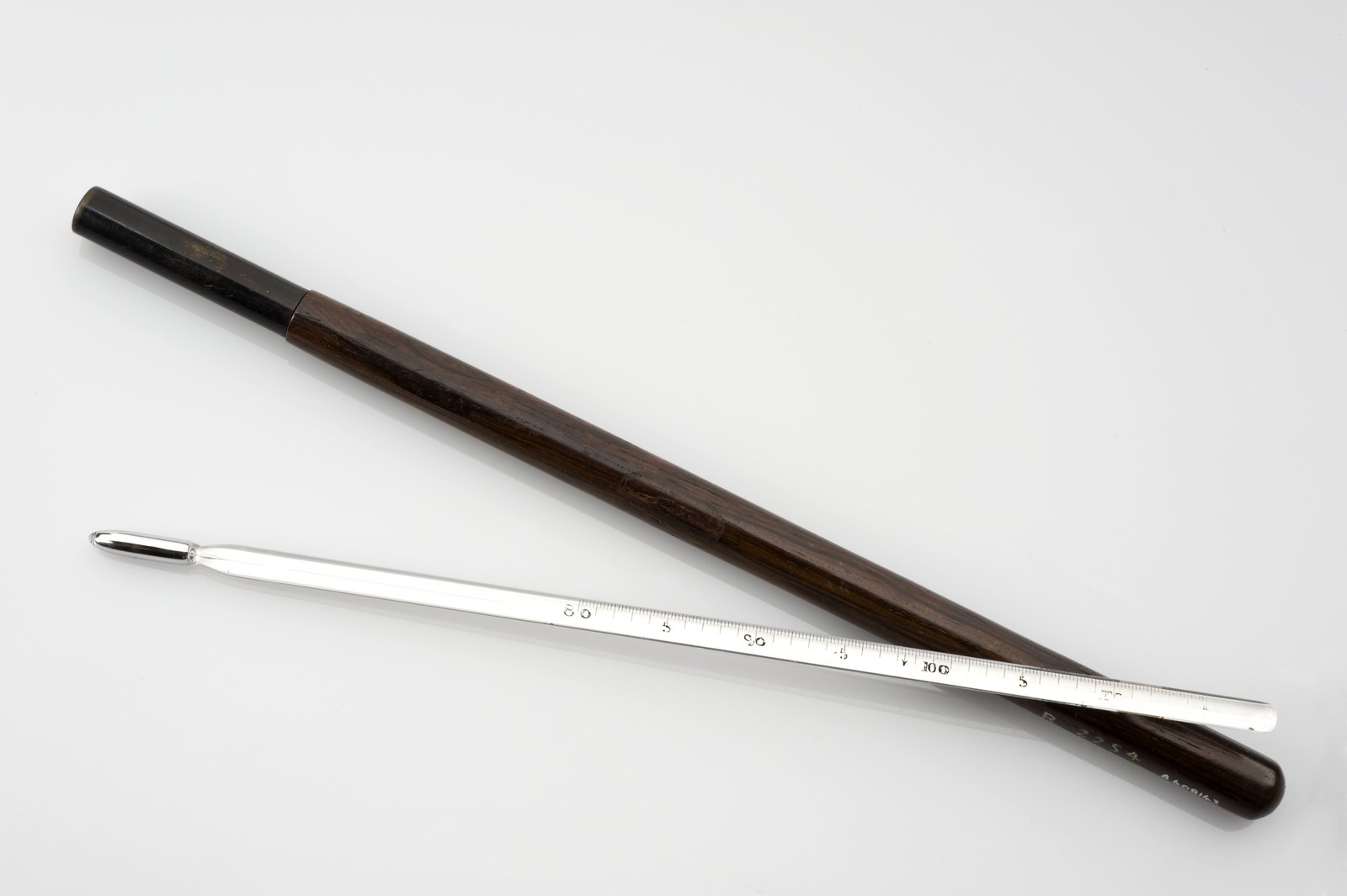 19th-century glass clinical thermometer next to its cylindrical dark wood case, both placed diagonally on a white background.