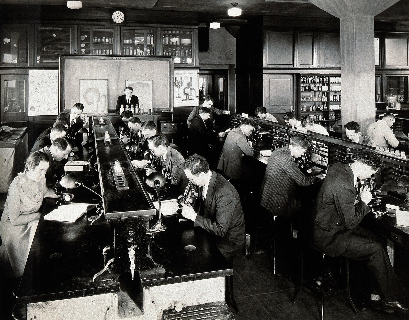 Schwarzweißfotografie von Studierenden, fast alle im Anzug und eine im hellen Kleid, die in Reihen an Labortischen sitzen und durch Mikroskope schauen, mit dem Professor vorne an einer Tafel, um 1933.
