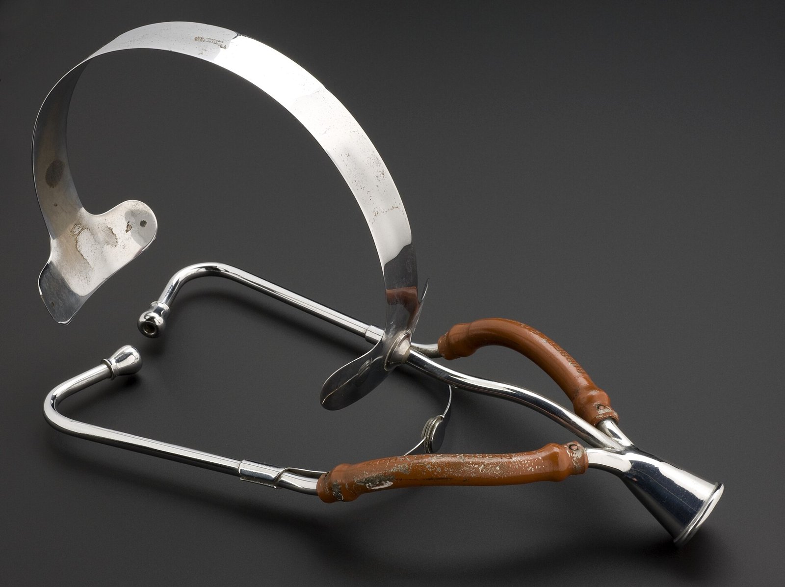 Estetoscopio obstétrico binaural de 1915 sobre fondo gris oscuro: dos varillas de metal plateado con tubos revestidos de cuero marrón gastado, una campana de metal en la punta y una banda metálica curva para apoyar en la cabeza.