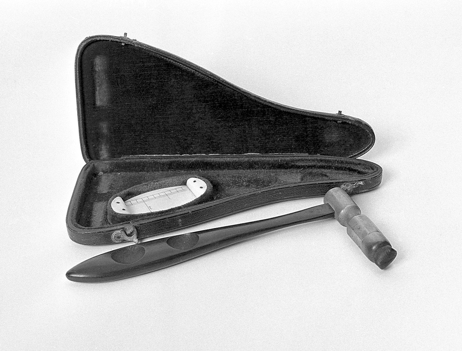 Fotografía en blanco y negro de un martillo de percusión antiguo, de mango oscuro y afilado con cabeza de metal, junto a un estuche abierto forrado de terciopelo que guarda una pequeña placa clara con escala, el plesímetro.
