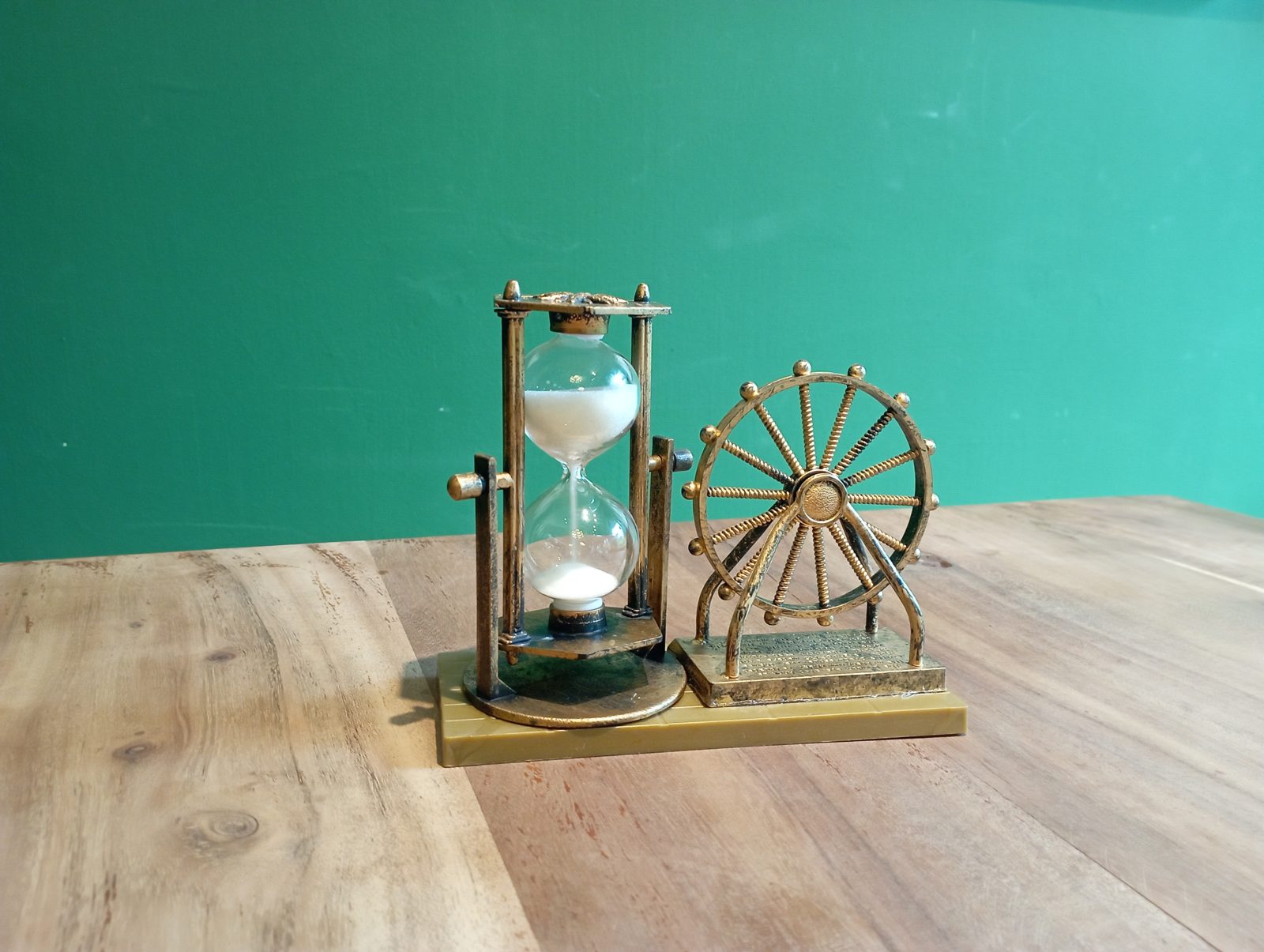 Eine Sanduhr aus Metall mit herabrinnendem weißem Sand, daneben ein kleines dekoratives Steuerrad, auf einem Holztisch vor einer grünen Wand.
