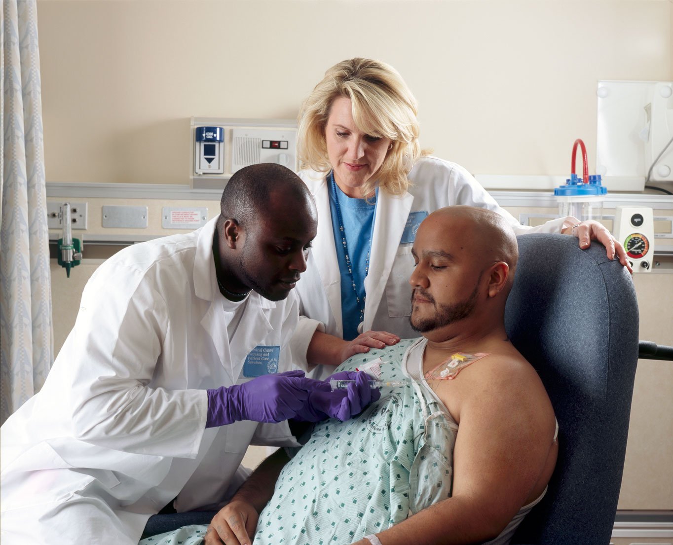A bald man with a beard, in a light green hospital gown, sits in a blue armchair while a professional in a white coat and purple gloves gives an injection through his chest catheter; behind them, a blonde female doctor in a white coat rests her hand on the patient's shoulder.