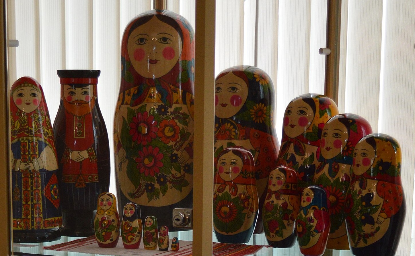 Bonecas de encaixe de madeira pintadas, de vários tamanhos, enfileiradas numa prateleira de vitrine, das grandes às bem pequenas