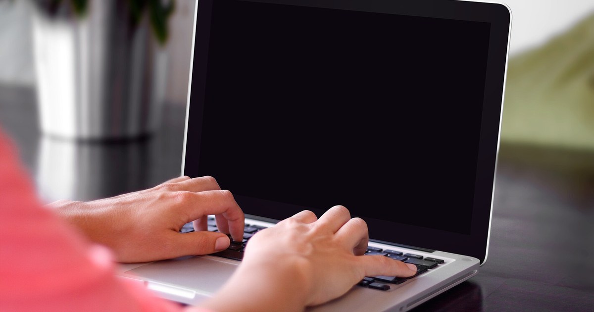 Hands typing on the keyboard of a silver laptop with the screen off, on a dark table, with the sleeve of a pink blouse in the foreground